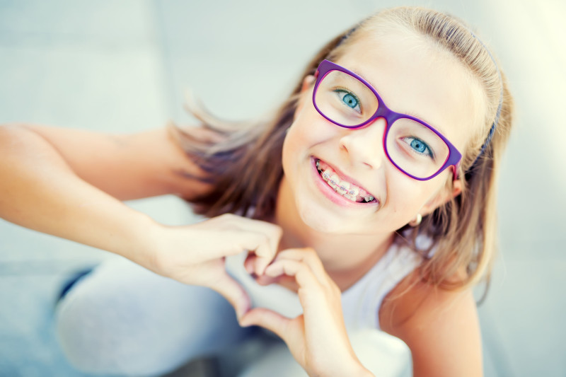Smiling little girl in with braces and glasses showing heart with hands.